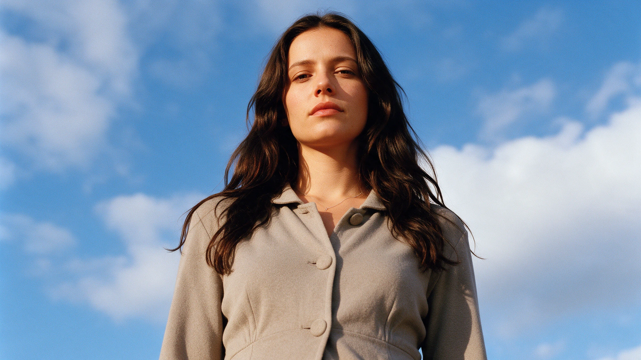 Woman in a beige coat gazing down at the camera beneath a blue, cloudy sky.