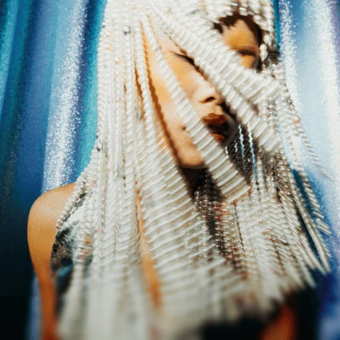 Person with an elaborate beaded headpiece, eyes closed, in front of a shimmering background.