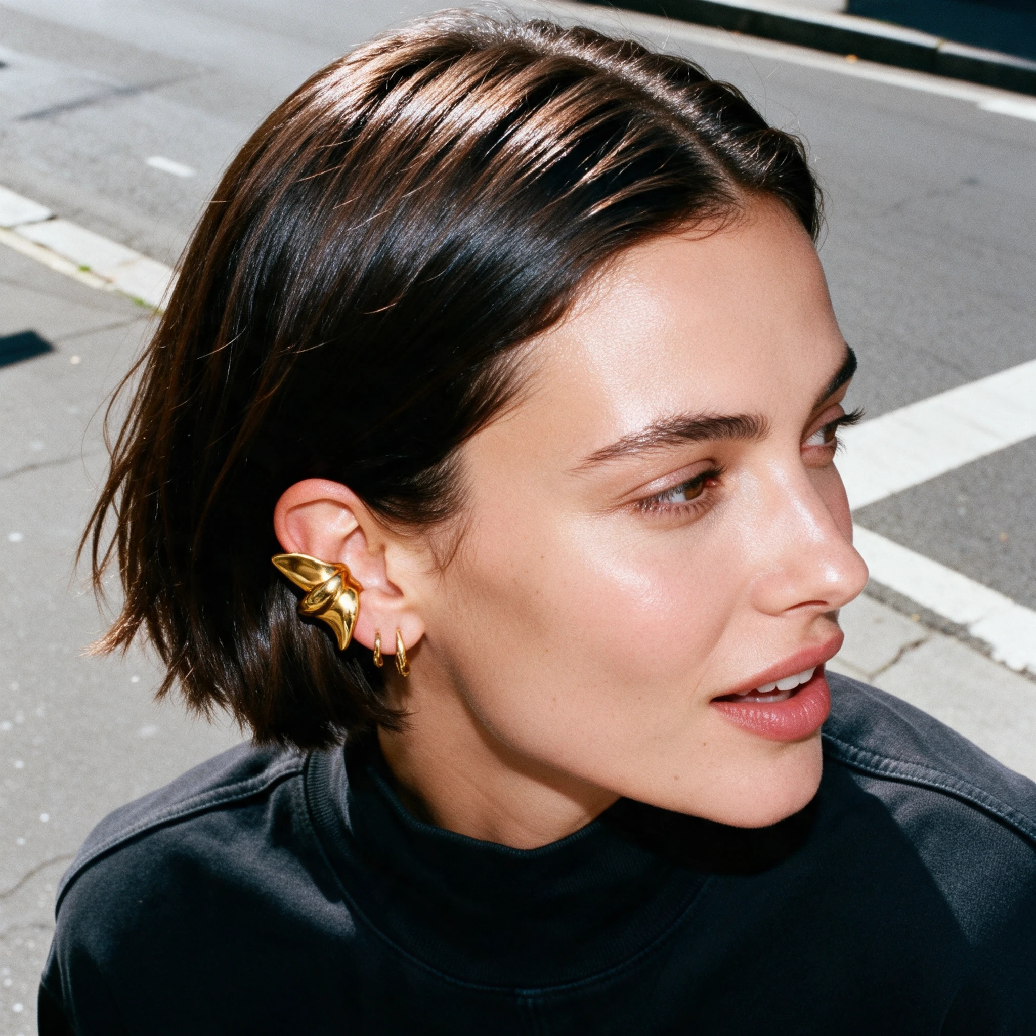 Close-up portrait of a person with short dark hair wearing gold ear jewelry outdoors.