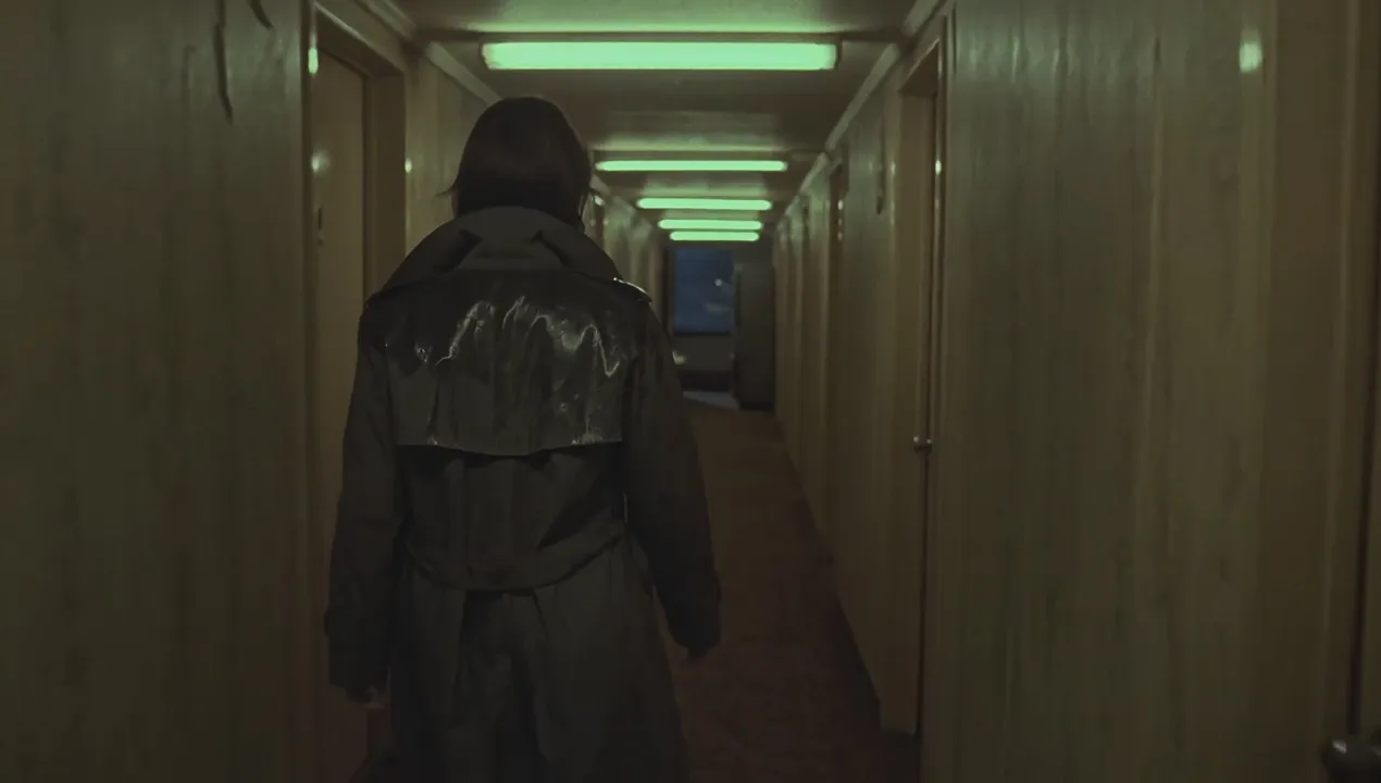 Slow handheld tracking shot behind a detective walking down a narrow motel hallway at midnight, her wet trench coat dripping a faint trail on the carpet. She passes three numbered doors, each closer than the last. As she passes the second door, the fluorescent tube above her flickers and dies, dropping her into shadow for a beat before the next tube buzzes on. She slows, fingertips grazing the peeling wallpaper, and glances at a room-service tray abandoned on the floor, a knocked-over glass still rocking slightly. Near the end of the hall, the vending machine rattles louder and its light stutters as she passes it. In the final seconds, the last door creaks open, spilling a slice of blue TV light across the floor. She freezes, one hand slipping into her coat pocket. The shot ends holding on her still silhouette against the light. Audio: muffled footsteps on damp carpet, a fluorescent buzz that cuts out then returns, the vending machine's uneven rattle growing closer, distant rain, one sharp door creak, a TV murmuring behind a wall, no music. 1970s neo-noir, 35mm film grain, warm green fluorescent cast mixed with cold blue spill from the open door, realistic motion. No readable text, no logos, no extra people on screen.