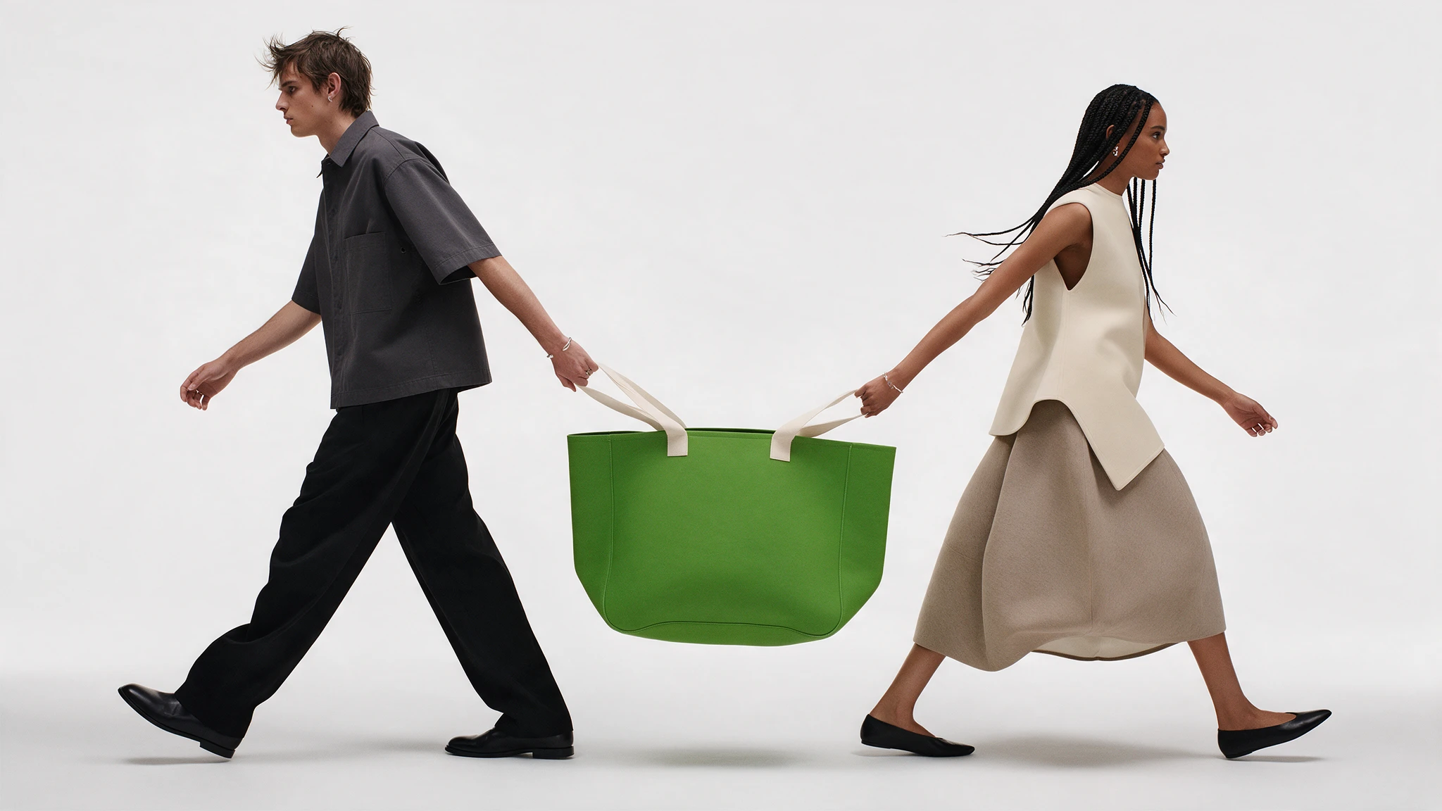 Two fashion models walk while carrying a large green tote bag between them.